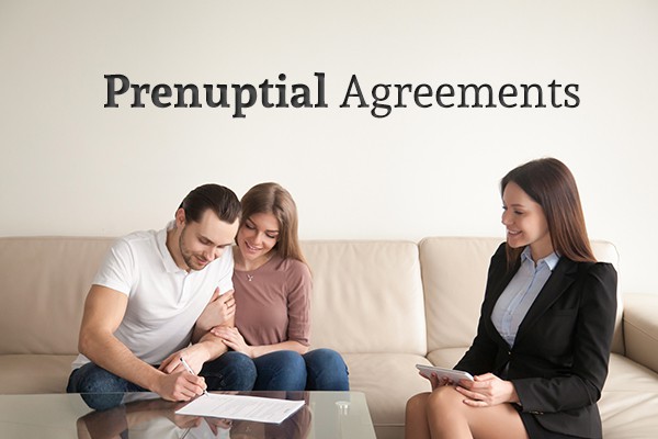 A happy couple sit on the couch signing a document across from a lawyer under the words "Prenuptial Agreements"